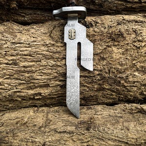 May include: A metal blade with a textured, mottled gray finish and a distinct cutout design along the upper edge. The blade features a small rectangular brass-colored inlay near the hilt, which is topped with a metallic guard and a dark handle.