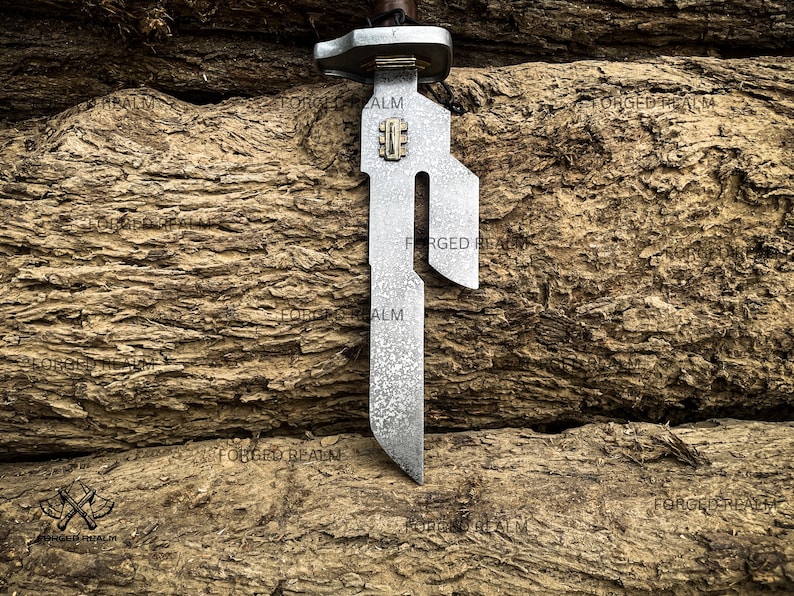May include: A metal blade with a textured, mottled gray finish and a distinct cutout design along the upper edge. The blade features a small rectangular brass-colored inlay near the hilt, which is topped with a metallic guard and a dark handle.