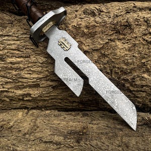 May include: A metal sword with a textured, mottled silver blade featuring a distinct cutout section and a gold-colored rectangular inlay. The hilt has a brown leather-wrapped handle and a silver crossguard with gold accents near the blade base.
