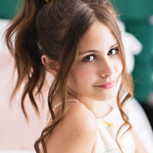 May include: A young person with long brown wavy hair pulled back in a ponytail with a yellow scrunchie. The person is wearing a white tank top with colorful abstract lines.