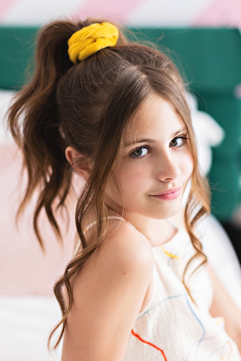 May include: A young person with long brown wavy hair pulled back in a ponytail with a yellow scrunchie. The person is wearing a white tank top with colorful abstract lines.