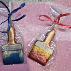 May include: Two paintbrush-shaped chocolate lollipops wrapped in clear plastic with blue and red ribbons. The lollipops are decorated with gold and silver paint.