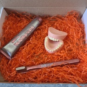 May include: A gift box filled with orange shredded paper containing a chocolate replica of a tube of toothpaste, a toothbrush, and a set of dentures.