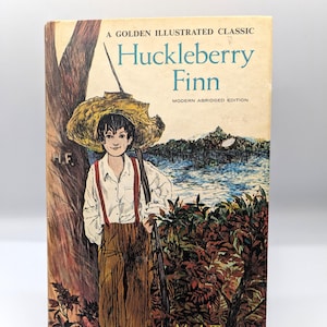 Könnte beinhalten: Ein Vintage-Exemplar von "Huckleberry Finn" von Mark Twain. Das Buchcover zeigt eine Illustration eines jungen Mannes mit Strohhut und Hosenträgern, der neben einem Baum steht. Der Titel ist blau, mit "A Golden Illustrated Classic" darüber.