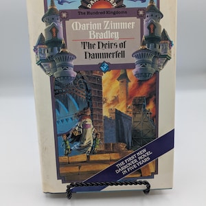 May include: A paperback book titled "The Heirs of Hammerfell" by Marion Zimmer Bradley. The cover art depicts a woman and children approaching a burning castle. The book has a blue and white cover with the text "Darkover" and "The Hundred Kingdoms".