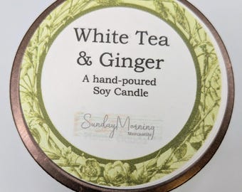 May include: A circular candle lid featuring the text "White Tea & Ginger" and "A hand-poured Soy Candle." The lid has a brown edge, a white central area, and a green floral border. The brand name "Sunday Morning Mercantile" is also visible.