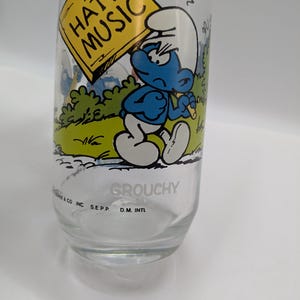 May include: A clear glass with a cartoon Smurf holding a sign saying "I HATE MUSIC." The Smurf is blue with a white hat, set against a green and white backdrop. The word "GROUCHY" is printed on the glass, a collectible item.