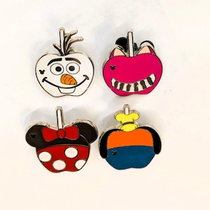 Op de afbeelding: Vier emaille pins in de vorm van appels. Eén pin heeft Olaf van Frozen, een andere heeft een Cheshire Cat-ontwerp, een derde heeft een Minnie Mouse-ontwerp en de laatste heeft een ontwerp met een koksmuts.