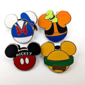 Può includere: Quattro spille smaltate raffiguranti i personaggi Disney: Paperino, Pippo, Topolino e Pluto. Ogni spilla ha la forma della testa di Topolino, con l'immagine del personaggio raffigurata in colori vivaci. Le spille sono su uno sfondo bianco.