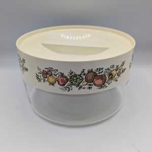 May include: A clear glass and cream-coloured plastic food storage container with a lid. The cream-coloured band around the container features a colourful fruit and vegetable design. The container is round and has a handle on the lid.
