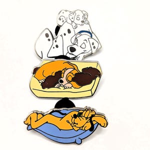 May include: Three enamel pins featuring cartoon dogs. One pin shows two dalmatians, another depicts a dog on a yellow bed, and the third shows a yellow dog on a blue bed. The pins have silver borders and are set against a white background.