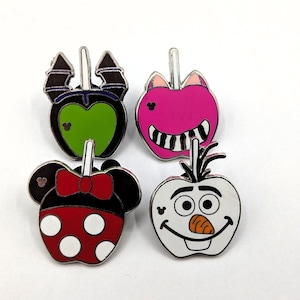 May include: Four metal enamel pins shaped like candy apples, each featuring a unique character design. One is black and green with horns, one is pink with a wide grin, one is red with white polka dots and a bow, and one is white with a carrot nose.