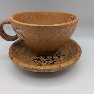 May include: A wooden teacup and saucer set. The cup has a handle and is decorated with black and white bamboo leaf designs. The saucer also features the same bamboo leaf design. The set is made of light brown wood.