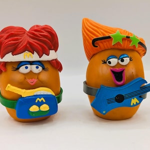 May include: Two vintage McDonald's Fry Kids figurines. One has red hair and a blue and yellow boombox. The other has orange hair and a blue guitar. Both have orange bodies, smiling faces, and green or gray bands.