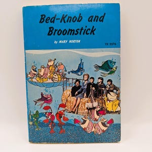 May include: A vintage paperback book titled "Bed-Knob and Broomstick" by Mary Norton. The cover features a blue background with illustrations of cartoon characters, fish, and people. The book has a retro design and is likely a children's book.