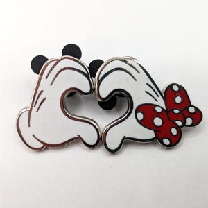 May include: A pair of enamel pins featuring white cartoon gloves forming a heart shape. One pin has a red bow with white polka dots. Black circular shapes are at the top of each pin.
