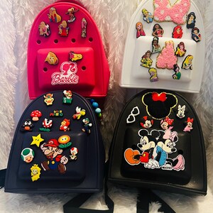 Puede incluir: Cuatro mini mochilas diferentes con varios diseños de personajes. Una mochila rosa con Barbie, una mochila blanca con princesas Disney, una mochila negra con Mickey Mouse y Minnie Mouse, y una mochila negra con Super Mario.