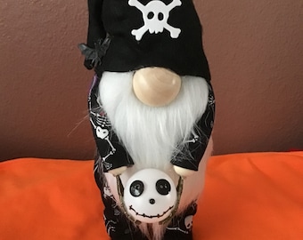 May include: A Halloween gnome wearing a black hat with a white skull and crossbones. The gnome has a white beard, a wooden nose, and holds a white Jack Skellington head. The gnome's outfit is black with white skeleton patterns.