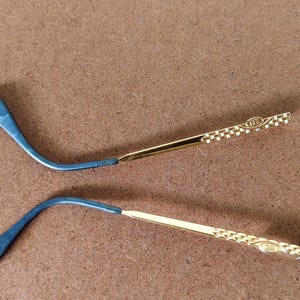 May include: Two eyeglass temples, one blue and one gold. The blue section curves and joins a gold arm with a decorative pattern. The gold arms have a small emblem at the end. The temples are set against a brown background.