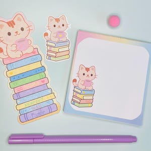 Peut inclure: Ensemble de papeterie avec un motif de chat de dessin animé. Comprend des autocollants, un bloc-notes, un stylo et un pompon rose. Le chat est représenté assis sur une pile de livres colorés et tenant une tasse.