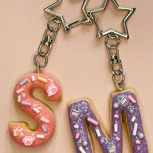 Könnte beinhalten: Zwei Schlüsselanhänger mit sternförmigen Anhängern. Einer hat einen rosa Buchstaben "S" mit Streuseln und Pfotenabdrücken. Der andere hat einen lila Buchstaben "M" mit Boba-Tee-Grafiken und Streuseln. Beide Buchstaben sind wie Kekse geformt.