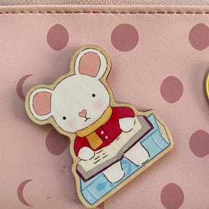 Könnte beinhalten: Ein hölzerner, mausförmiger Anstecker mit roter Jacke und gelbem Schal, der ein Buch liest. Der Anstecker befindet sich auf einem rosa Hintergrund mit Punkten und einem Reißverschluss.