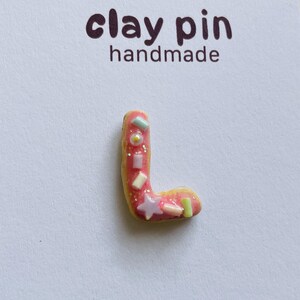 Könnte beinhalten: Handgemachter Ton-Pin in Form des Buchstabens "L". Der Pin ist rosa mit gelben Rändern und mit pastellfarbenen Streuseln und einem Stern verziert. Der Text "clay pin handmade" ist über dem Pin aufgedruckt.