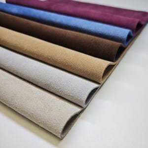 Op de afbeelding: Een close-up van een stapel suède stofstalen in verschillende kleuren. De kleuren zijn bordeauxrood, blauw, bruin, beige, grijs en lichtbeige. De stof heeft een zachte, getextureerde uitstraling.