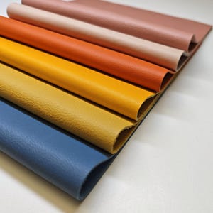 May include: A collection of colorful leather swatches in shades of blue, yellow, orange, and pink. The leather has a textured surface and is arranged in a layered fan shape, showcasing the variety of colors and textures available.
