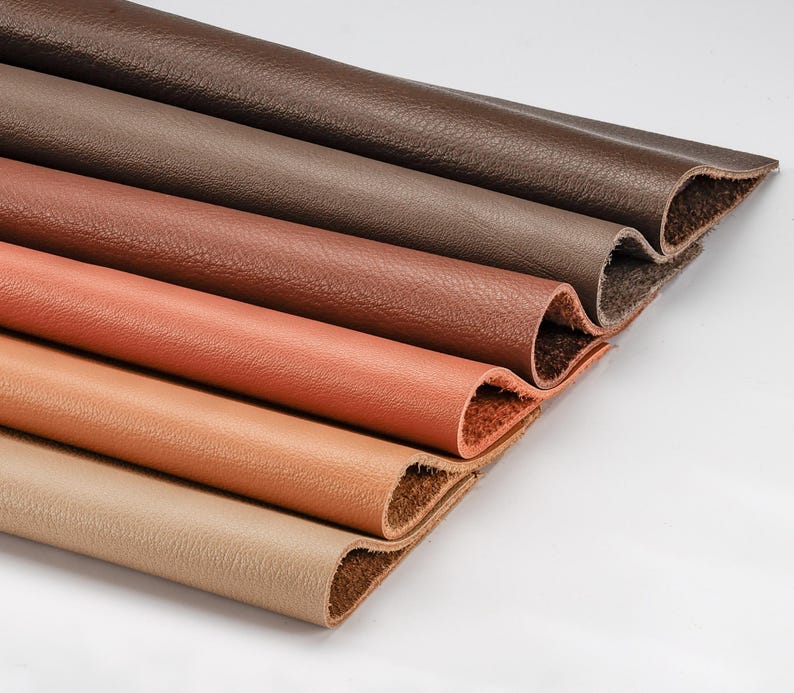 May include: Close-up of several rolled pieces of leather in varying shades of brown, from dark chocolate to tan. The edges of the leather are visible, showing the texture and color variations. The leather appears to be new and unused.