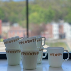 May include: Set of six white ceramic mugs with a brown floral pattern around the rim. The mugs are stacked and arranged on a windowsill with a blurry view of trees and houses in the background.