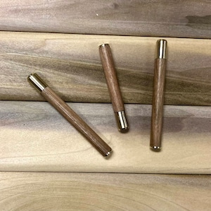 May include: Three wooden pens with gold-colored metal caps. The pens are arranged in a triangular pattern on a wooden surface.