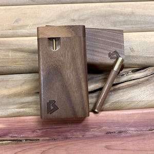 May include: A wooden box with a brass lid and a small wooden pipe. The box is made of walnut wood and has a dark brown finish. The pipe is made of wood and has a brass tip. The box and pipe are both engraved with a logo.