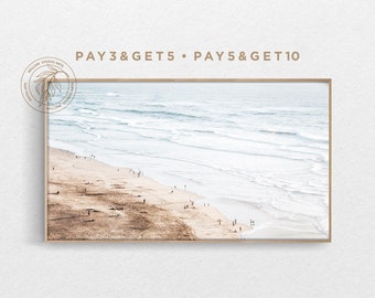 May include: An aerial view of a sandy beach with ocean waves crashing on the shore. The beach is sparsely populated with people walking along the shoreline. The image is framed in a light brown wood frame.