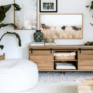 May include: A wooden media console with sliding doors, featuring a white knitted pouf, a vase with a geometric pattern, and framed artwork with images of a surfboard, a group of people, and a herd of wildebeest. The console is decorated with books, a woven basket, and a white blanket.
