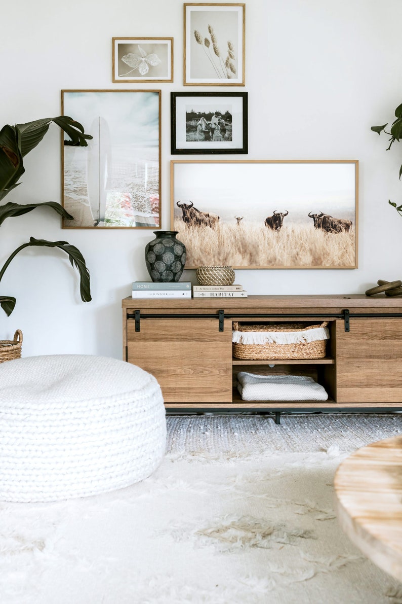May include: A wooden media console with sliding doors, featuring a white knitted pouf, a vase with a geometric pattern, and framed artwork with images of a surfboard, a group of people, and a herd of wildebeest. The console is decorated with books, a woven basket, and a white blanket.