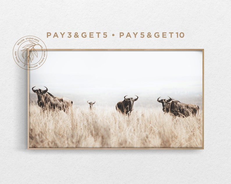 May include: A framed photograph of four wildebeest standing in a field of tall, dry grass. The background is a hazy, light blue sky. The image is framed in a light brown wood frame.