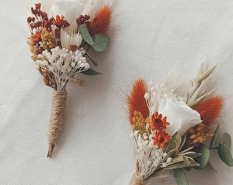 Peut inclure: Deux boutonnières composées de roses blanches, d'herbes séchées orange, de petites fleurs blanches et de feuilles d'eucalyptus vertes. Les tiges sont enveloppées de ficelle de jute naturelle, offrant un aspect rustique aux tons blanc, orange et vert.