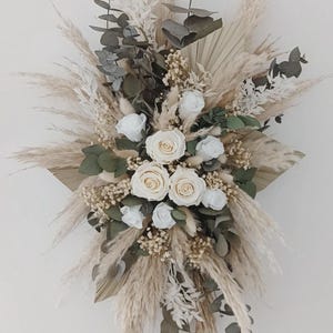 Peut inclure: Composition florale décorative avec des roses blanches, de l'herbe de la pampa et des feuilles séchées. L'arrangement présente des tons beige, crème et vert, créant une esthétique naturelle et élégante. Idéal pour la décoration intérieure ou les événements.