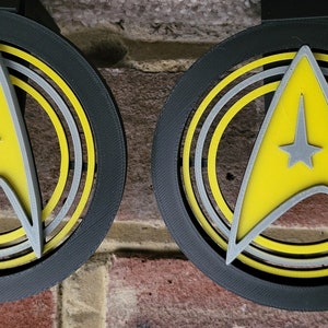 May include: Two black circular ornaments with a yellow Starfleet insignia in the center. The insignia is surrounded by concentric circles in yellow and gray.