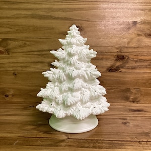May include: A matte white ceramic Christmas tree figurine featuring a textured, needle-like surface and small circular openings throughout the branches, set upon a smooth, circular base. The structure is conical and mimics the appearance of a pine tree.