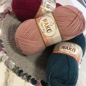 May include: Three balls of yarn in different colours: burgundy, light pink, and teal. Each ball has a label that says "NAKO Super Inci" in gold lettering.