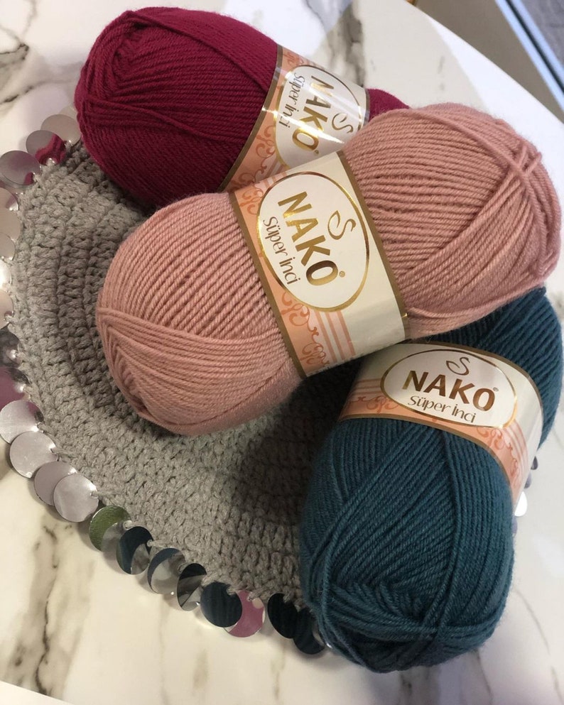 May include: Three balls of yarn in different colours: burgundy, light pink, and teal. Each ball has a label that says "NAKO Super Inci" in gold lettering.