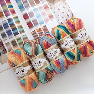 Pode incluir: Seis novelos coloridos de fio Alize Diva, cada um com uma faixa de papel. O fio é uma mistura de cores vibrantes, incluindo laranja, amarelo, verde, azul e roxo. Um gráfico de cores com várias amostras é visível no fundo.