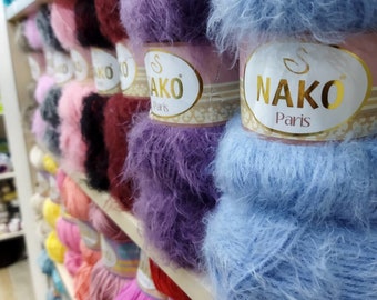 Op de afbeelding: Een close-up van een plank met verschillende bollen garen in verschillende kleuren, waaronder roze, paars, blauw en rood. Het garen is geëtiketteerd "Nako Paris" en is gemaakt van een zacht, pluche materiaal.