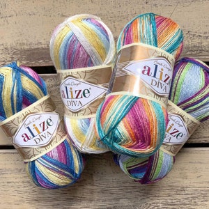 Pode incluir: Vários novelos de fio Alize Diva em várias cores, incluindo azul, amarelo, rosa e verde. Cada novelo é embrulhado com um rótulo que diz "Alize Diva Batik Design". O fio está disposto sobre uma superfície de madeira.