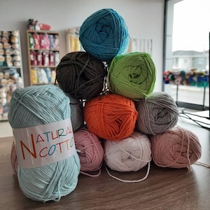 Pode incluir: Uma pilha de oito novelos de lã em várias cores, incluindo turquesa, marrom, verde, laranja, cinza, branco e rosa. O novelo de lã de baixo é azul claro e tem o texto "NATURAL COTTO" impresso no rótulo.