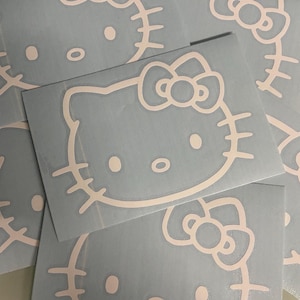 Puede incluir: Calcoman&iacute;as de la cara de Hello Kitty en rosa p&aacute;lido sobre un fondo azul claro. Las calcoman&iacute;as presentan la ic&oacute;nica cara del gato con un lazo, ojos, nariz y bigotes. Las calcoman&iacute;as est&aacute;n recortadas y listas para aplicar. Varias calcoman&iacute;as est&aacute;n apiladas.