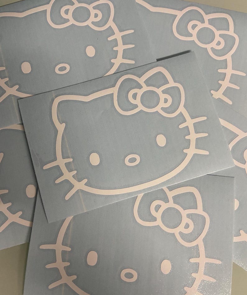 Puede incluir: Calcoman&iacute;as de la cara de Hello Kitty en rosa p&aacute;lido sobre un fondo azul claro. Las calcoman&iacute;as presentan la ic&oacute;nica cara del gato con un lazo, ojos, nariz y bigotes. Las calcoman&iacute;as est&aacute;n recortadas y listas para aplicar. Varias calcoman&iacute;as est&aacute;n apiladas.