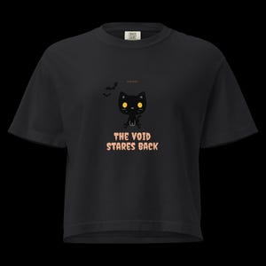 May include: A black short-sleeved t-shirt featuring a graphic of a small black cat with yellow eyes and two bats. Below the cat, the text reads THE VOID STARES BACK in a light orange font. The shirt has a crew neckline and a standard fit.